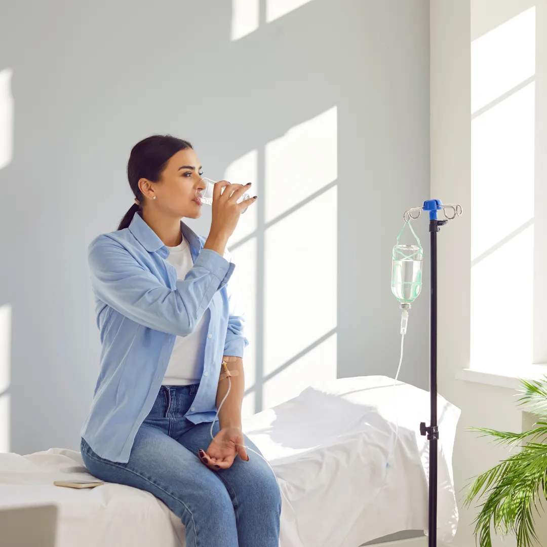 A young woman seated comfortably while receiving IV therapy in a bright, sunlit room, sipping water as part of a rejuvenation or wellness session.