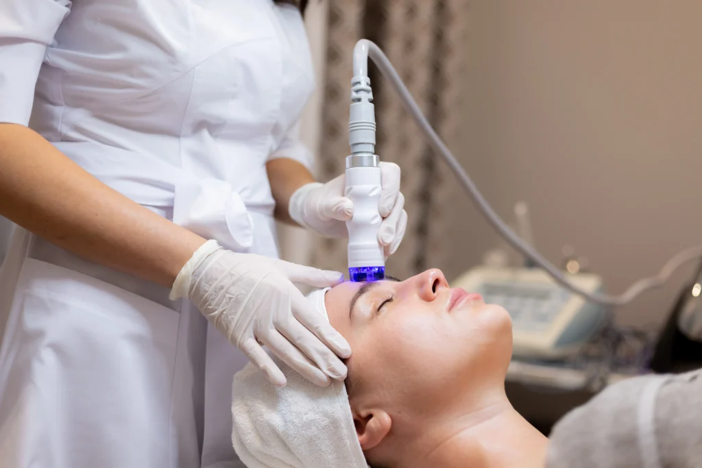 Close-up of a woman undergoing a hydrafacial procedure at an aesthetic clinic. A gloved aesthetician is using a modern skin treatment device with blue light technology on the client's forehead, demonstrating professional care and advanced facial rejuvenation techniques.
